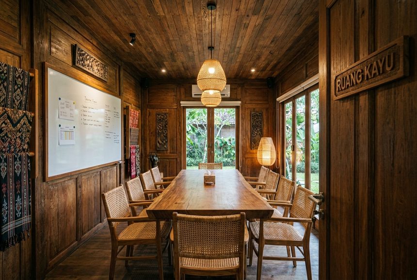 Ruang rapat berdinding kayu dengan meja panjang, dua belas kursi rotan, dan papan tulis putih.