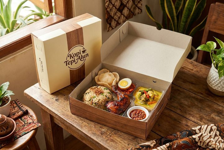 Kotak nasi kertas berisi nasi goreng, ayam bakar, telur, dan sayur, di sebelah kotak tertutup berlogo kafe.